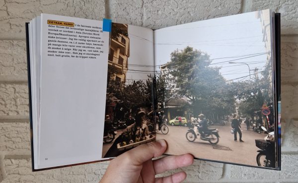 En bok holdes åpen på en side som viser et bilde av en gatescene i Hanoi. Fokuset i bildet er på en taxi som er ganske langt fra fotografen, den kvinnelige sjåføren poserer foran taxien. I forgrunnen i bildet er det variert trafikk av fotgjengere, sykler og mopeder, disse er ikke i fokus og derfor varierende grad av uklare.